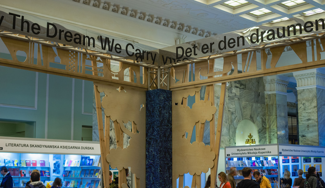 Part of a wooden stand at a book fair. At the top it says "The dream we carry" and "Det er den draumen". Small stands of polish publishers in the background. 